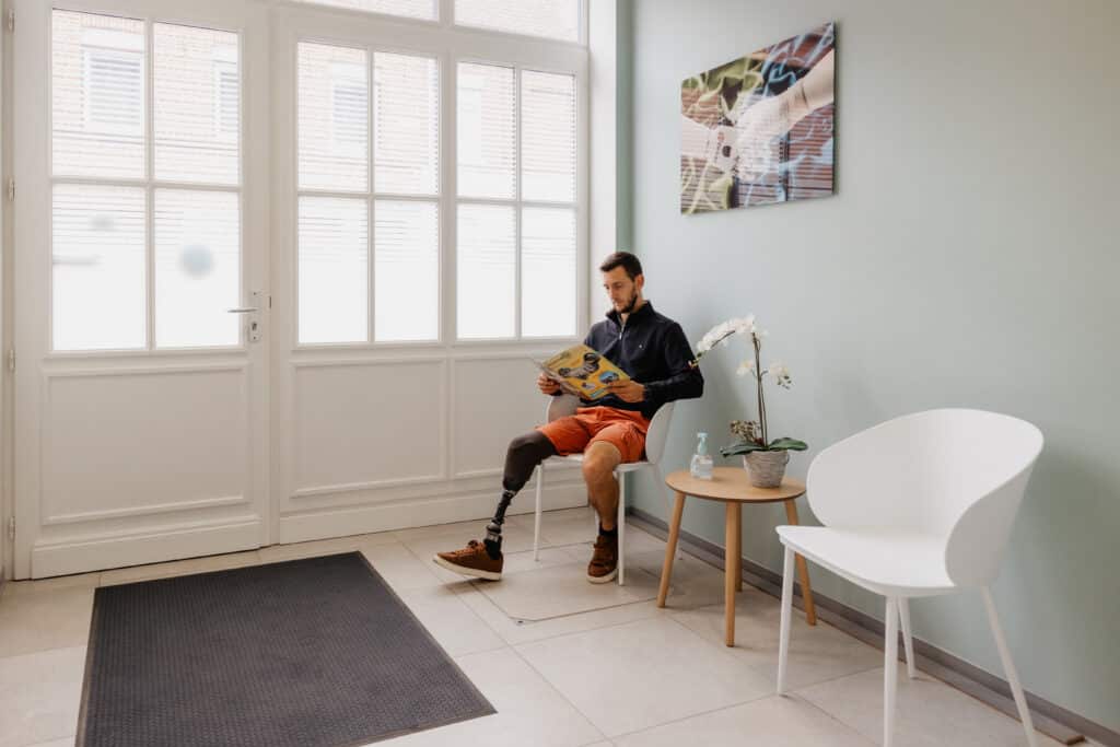 Patient avec prothèse lisant un magazine dans une salle d'attente moderne. Décoration épurée, tableau bionique sur le mur vert clair.