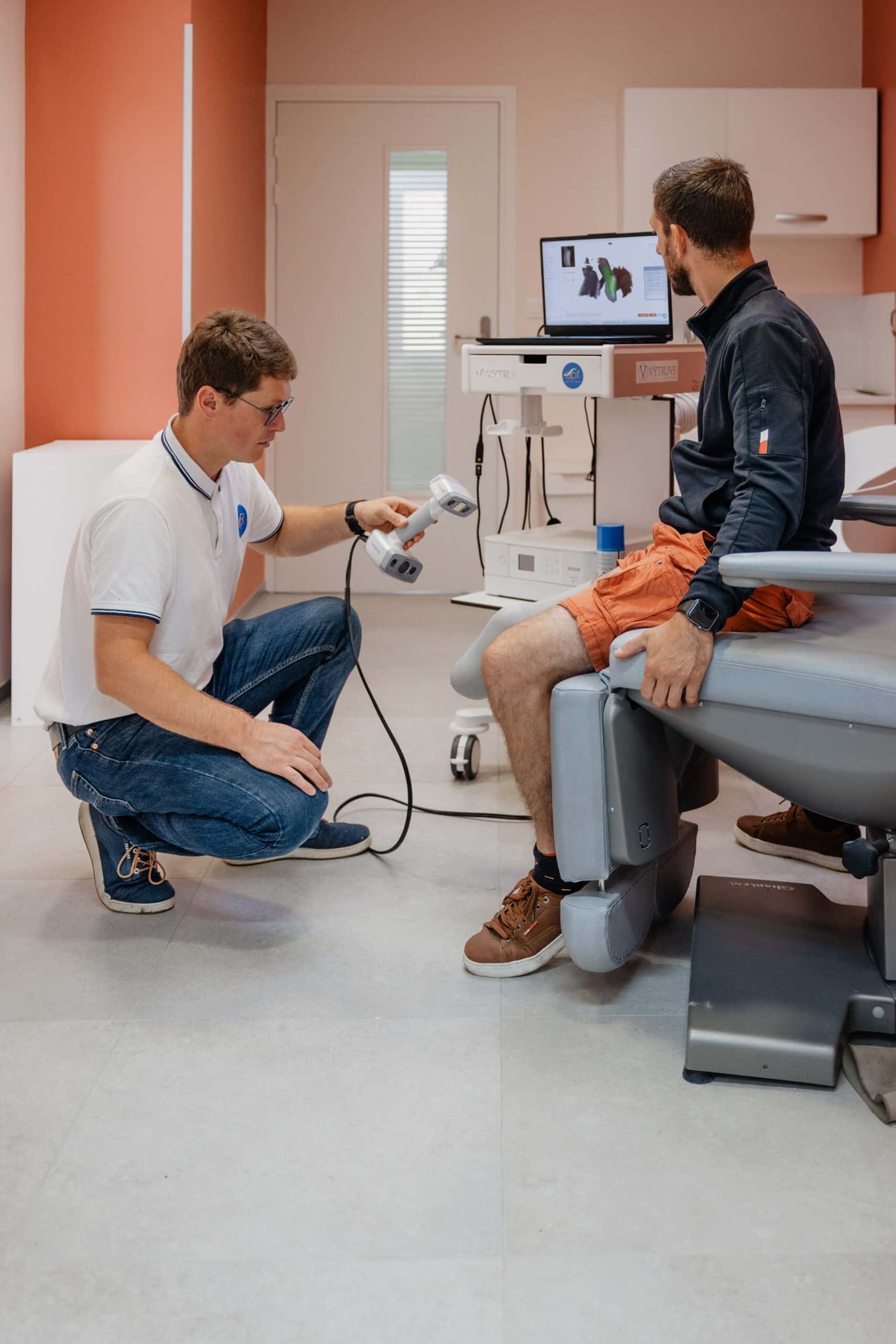 Professionnel de santé utilise un scanner 3D (Peel 3D) pour la mesure numérique de la jambe d'un patient assis. Écran Vytruve visible.